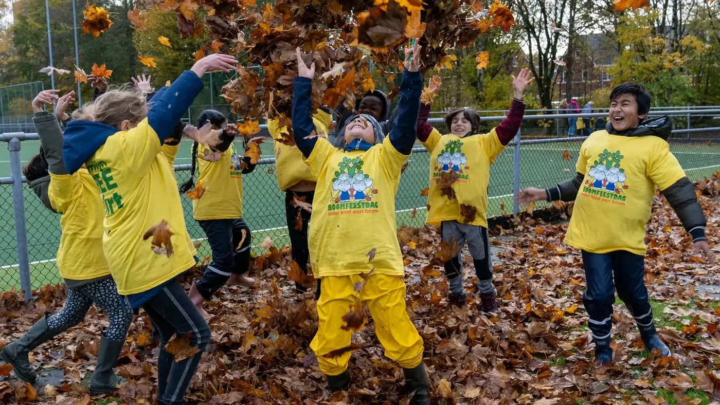 Kinderen in gele Boomfeestdag-shirts gooien handenvol herfstbladeren de lucht in.