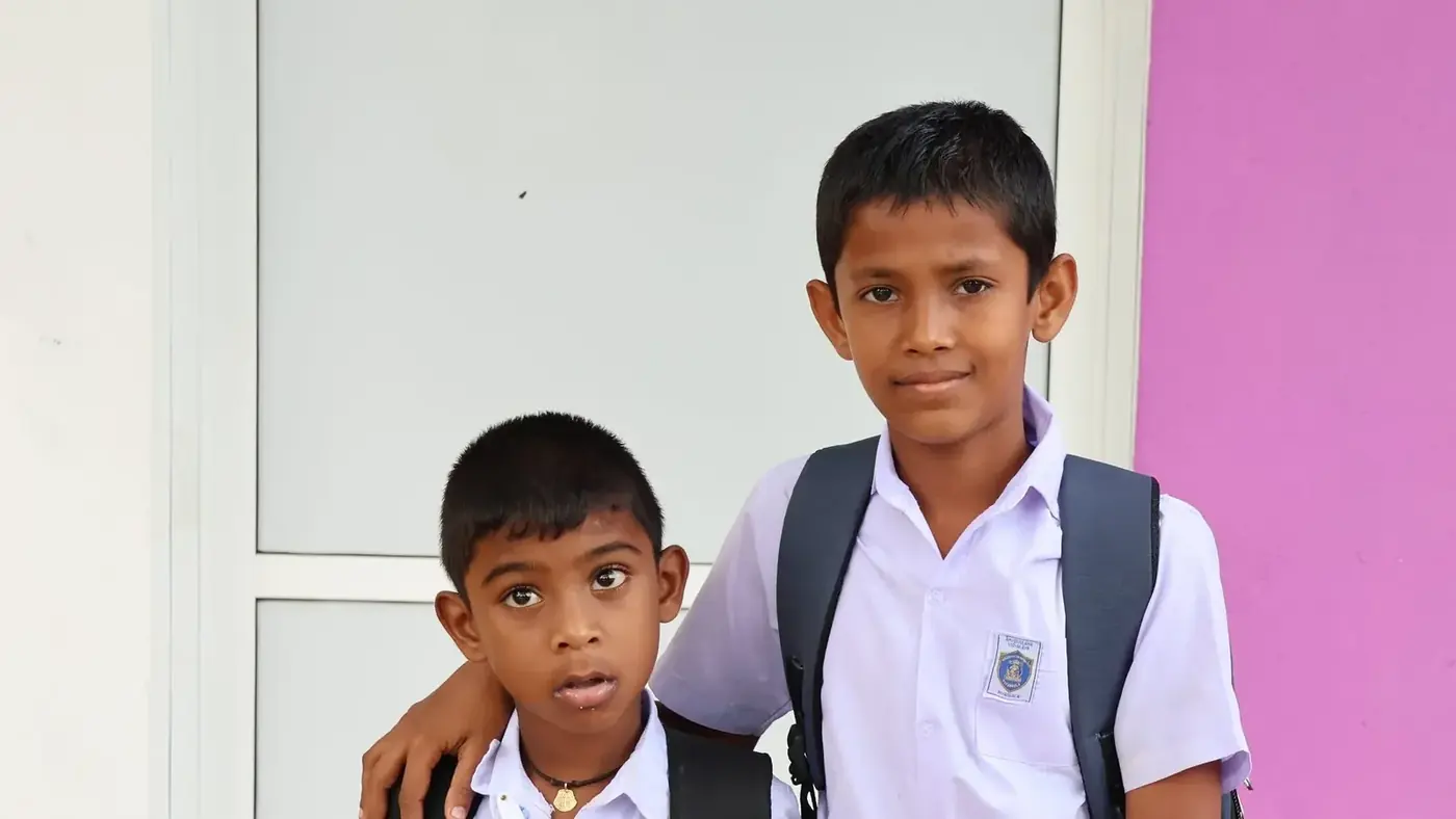 Twee jongens in wit schooluniform staan naast elkaar voor een schoolgebouw; de oudste jongen heeft zijn arm om de jongste heen.