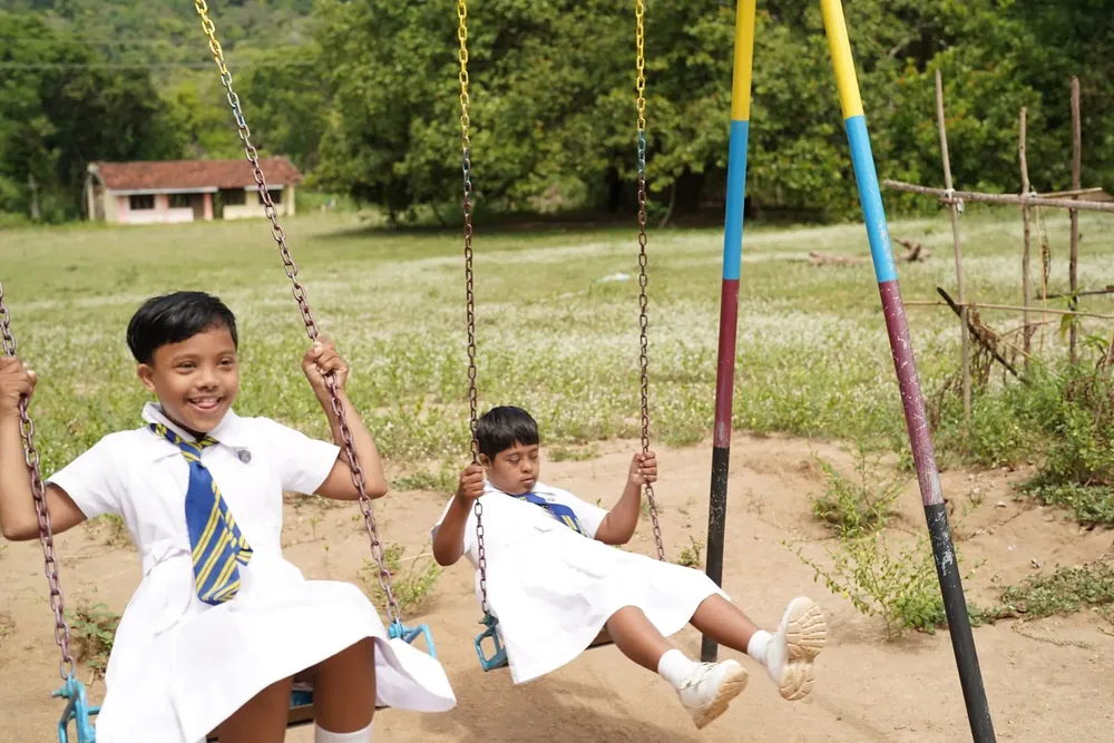 Twee meisjes in schooluniform schommelen naast elkaar op een schoolplein in Sri Lanka.