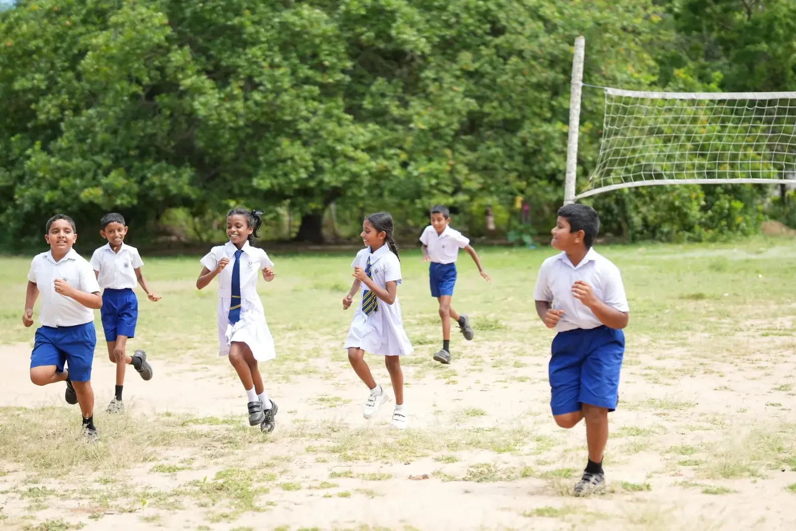 Zes kinderen in schooluniform rennen lachend over een zandveld naast een volleybalnet.